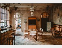 Ein rustikal eingerichteter Raum mit Holztisch und mehreren Holzstühlen in der Mitte, umgeben von Wänden in Betonoptik. Im Hintergrund sind zwei bogenförmige Türen, eine Lampe mit mehreren weißen Leuchten an der Decke und verschiedene Vintage-Möbel sowie Dekorationselemente sichtbar.