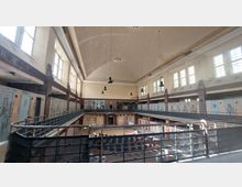 Innenansicht eines großen historischen Gebäudes mit hohen Decken und Oberlichtern. Der Raum hat eine umlaufende Galerie mit Geländern, alten Türen und Fenstern, und die Wände zeigen Spuren von Verfall.