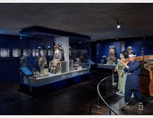 Ausstellungsraum eines Museums mit Vitrinen, in denen traditionelle japanische Samurai-Rüstungen präsentiert werden. Im Vordergrund stehen lebensgroße Figuren in historischen Kleidern, die ein japanisches Szenerie-Element darstellen, um die Exponate zu ergänzen.