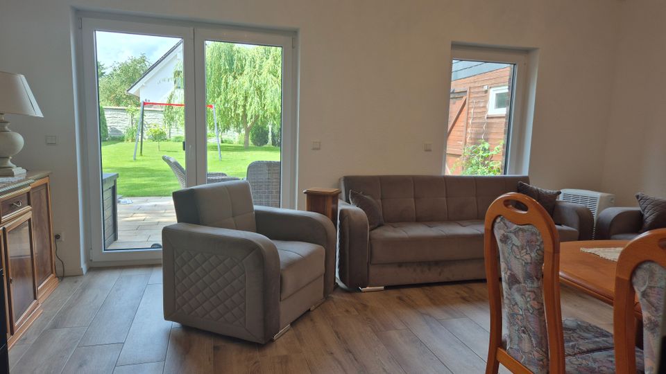 Helles Wohnzimmer mit Holzfußboden, grauem Sessel und Sofa sowie einem angrenzenden Essbereich. Große Terrassentür und ein Seitenfenster bieten Blick in den Garten mit Rasen und Terrasse.