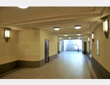 Ein unterirdischer Durchgang mit beigen Wänden und einer hellen Decke, beleuchtet durch mehrere Wand- und Deckenleuchten. Am Ende des Flurs befinden sich Treppen, die nach oben führen, sowie eine metallene Gittertür.