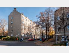 Straßenecke in einer Wohngegend mit mehrstöckigen Wohngebäuden aus Putz, teils mit Graffiti an der Fassade. Die Straße ist von kahlen Bäumen gesäumt, herbstliches Laub liegt auf dem Gehweg; geparkte Autos und ein Fahrradfahrer sind sichtbar.