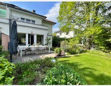 Zweistöckiges Wohnhaus mit weißer Fassade und Holzterrasse im Garten. Im Vordergrund ist ein gepflegter Garten mit grüner Wiese, Blumenbeeten und großen Bäumen sichtbar.