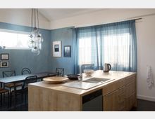 Eine moderne Küche mit Kochinsel aus Holz, Spüle und Wasserhahn im Vordergrund. Im Hintergrund befinden sich ein Esstisch mit schwarzen Stühlen, blaue Vorhänge an großen Fenstern und eine blaue Wand mit gerahmten Bildern.