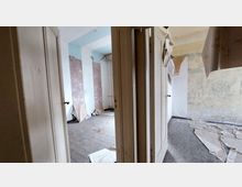 Ein verlassenes, heruntergekommenes Zimmer mit abgeblätterter Tapete, freiliegendem Bodenbelag und einer offenen Tür, die zu einem angrenzenden Raum führt. Auf dem Boden liegen Reste von Tapeten und Schutt, während die Wände mit verblassten und teils beschädigten Farben bedeckt sind.