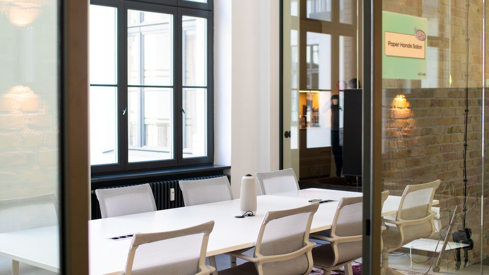 Moderner Besprechungsraum hinter Glaswänden mit langem weißen Konferenztisch und mehreren hellen Drehstühlen auf einem gemusterten Teppich. Große Sprossenfenster und eine Ziegelwand prägen den hellen Raum.