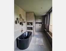 Modernes Badezimmer mit einer freistehenden schwarzen Badewanne, Wänden und Boden aus grauen Fliesen sowie einem großen Fenster, das Tageslicht hereinlässt. An der Wand hängt ein Dekorationsobjekt aus runden Spiegeln, und eingebaute Nischen bieten Stauraum für Accessoires.