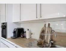 Moderne Küche mit weißen Hochglanzschränken, einem Holzarbeitsplatz und einer Edelstahlspüle. Auf der Arbeitsfläche befinden sich eine Kaffeemaschine, Schneidebretter, ein Wasserkocher sowie eine Teekanne mit grünem Tee.