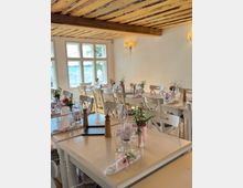 Ein gemütlich eingerichteter Speiseraum mit hellen Holztischen und -stühlen, dekoriert mit Blumenvasen, Servietten und Gläsern; ein Holzbalken an der Decke und große Fenster mit Blick auf Wasser im Hintergrund.