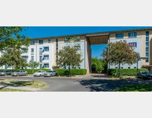 Mehrstöckiges Wohngebäude mit weißer und beigefarbener Fassade, großen Fenstern und einer Durchfahrt in der Mitte. Im Vordergrund stehen blühende Bäume, geparkte Autos und ein asphaltierter Weg.