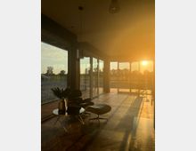 Modernes Wohnzimmer mit großen bodentiefen Glasfenstern, durch die die Abendsonne hereinstrahlt. Im Vordergrund stehen ein schwarzer Sessel mit passendem Hocker sowie ein runder Beistelltisch mit einer Vase, eingerichtet auf einem Holzparkettboden.