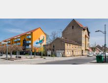 Straßenansicht mit einem großen gelben Wohngebäude, das an der Seite ein buntes Wandgemälde zeigt, sowie mehreren kleineren, älteren Gebäuden mit verblassender Fassade. Im Vordergrund sind eine gepflasterte Fläche, junge Bäume und parkende Autos zu sehen, die an einer ruhigen Straße liegen.