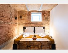 Schlafzimmer mit freiliegenden Ziegelwänden und gewölbter Decke. Im Raum stehen ein Doppelbett mit weißer Bettwäsche und gelber Decke, flankiert von zwei Nachtlampen; im Vordergrund befindet sich eine rustikale Holztruhe mit einer Schale kleiner Pflanzen.