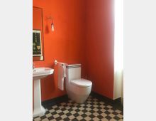 Badezimmer mit orange gestrichenen Wänden, einem weißen Waschbecken mit Spiegel, einer Toilettenschüssel und schwarz-weiß karierten Fliesen auf dem Boden. Eine einfache Glühbirne hängt an der Wand, rechts fällt Tageslicht durch einen Vorhang.