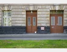 Fassade eines historischen Gebäudes mit hellem Steinmauerwerk und einer unteren schwarzen Steinverkleidung. Zwei braune Holztüren mit verzierten Gitterelementen sind nebeneinander angeordnet, daneben ein vergittertes Fenster; im Vordergrund eine kleine Rasenfläche und eine Straße.