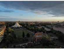 Luftaufnahme einer urbanen Umgebung mit dem Tempodrom, einem weißen, zeltartigen Veranstaltungsgebäude, umgeben von Bäumen und einem Fußballfeld im Vordergrund. Im Hintergrund ist die Stadtlandschaft von Berlin mit historischen Gebäuden, modernen Strukturen und Kirchen zu sehen, unter einem teils bewölkten Himmel.