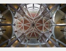 Innenansicht einer Kirche mit einer kunstvoll verzierten, sternförmigen Decke in Rosa- und Goldtönen, getragen von hohen Säulen. Rundbögen mit filigranen Geländern umgeben den Raum.