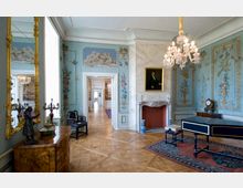 Elegant gestalteter historischer Salon mit kunstvoll bemalter Wandvertäfelung, einem Kamin mit Marmorumrahmung, einem Kronleuchter sowie antiken Möbeln wie einem Klavier und einer Kommode mit dekorativen Figuren. Durch eine geöffnete Tür sind weitere prunkvolle Räume sichtbar.