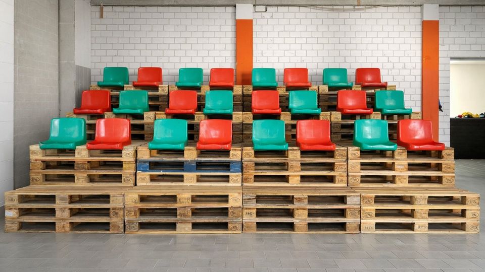 Provisorische Tribüne in einer Industriehalle: bunte Kunststoffsitze in Rot und Grün stehen in mehreren Reihen auf gestapelten Holzpaletten. Im Hintergrund eine weiße Ziegelwand mit orangefarbenen Stützen und grauer Beton­decke, davor ein gefliester Boden.