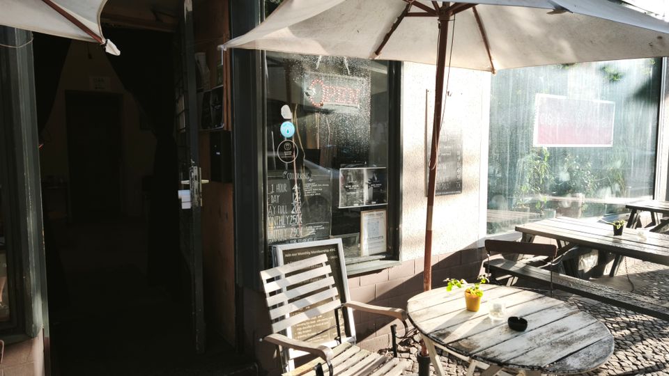 Außenbereich eines kleinen Cafés mit runden Holztischen und Stühlen unter großen Sonnenschirmen auf Kopfsteinpflaster. Dahinter stehen eine Glasfront mit Tür und Fenster sowie eine offene Eingangstür in den Innenraum.