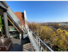 Dachterrasse einer Wohnung mit Geländer, Topfpflanzen und einem Sessel im Vordergrund. Im Hintergrund Blick auf herbstliche Bäume und eine Stadtlandschaft unter klarem, blauem Himmel.