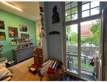 Ein gemütliches Zimmer mit grüner Wand, dekoriert mit Tierfotografien und einer grauen Kommode. Rechts befinden sich große Fenster und eine Balkontür, die zu einem kleinen begrünten Balkon mit Tisch und Stühlen führt.