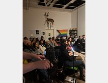 Indoor event space with rows of chairs facing a small presentation area, set in a white-walled room with exposed ceiling beams and track lighting. Wall decor includes a mounted antelope sculpture above a door, several posters, and a large pride flag hanging beside metal shelving.