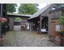 Hof mit Kopfsteinpflaster und rustikalen Holzbauten, darunter ein Holzschuppen mit Vordach, dekoriert mit Tischen und Pflanzen; im Hintergrund sind weitere Gebäude und Bäume zu sehen.