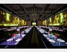 Großer Veranstaltungssaal mit langen Banketttischen, die mit weißen Tellern, Gläsern und roten Blumen dekoriert sind. Die Wände zeigen großflächige beleuchtete Projektionen mit historischen Motiven, während moderne Lichttechnik an der Decke angebracht ist.