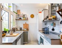 Helle, moderne Küche mit weißen Schränken und grauen Arbeitsplatten, ausgestattet mit einem Cerankochfeld, einem Ofen und einem Spülbecken. Die Wände sind mit weißen Fliesen verkleidet, Regale mit Küchenutensilien und Pflanzen schmücken den Raum, während große Fenster für viel Tageslicht sorgen.