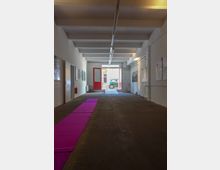 Langer Korridor in einem minimalistischen Gebäude mit weiß gestrichenen Wänden, Betondecke und dunklem Bodenbelag. Ein pinker Teppich führt zu einer roten Tür im Hintergrund, durch die Tageslicht einfällt, und es sind gerahmte Bilder an den Wänden aufgehängt.