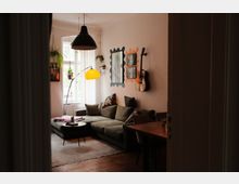 Gemütliches Wohnzimmer mit großer Eckcouch, kleinem runden Couchtisch und Stehlampe neben einem hohen Fenster mit hängenden Pflanzen. An der Wand hängen dekorative Spiegel und eine Gitarre, dazu eine große Pendelleuchte über dem Sitzbereich.