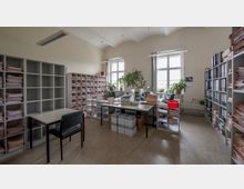 Ein Büro mit hohen Decken, großen Fenstern und mehreren Regalen, die mit Aktenordnern und Papierstapeln gefüllt sind. In der Raummitte stehen Schreibtische mit Büromaterialien, Druckern und Pflanzen, umgeben von Archivordnern und Kartons.