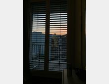 Fenster mit heruntergelassenen Jalousien, durch die ein Blick auf eine Stadtlandschaft mit modernen Gebäuden und einem leicht gefärbten Abendhimmel erkennbar ist. Im Vordergrund sind Teile eines kleinen Innenraums sichtbar.