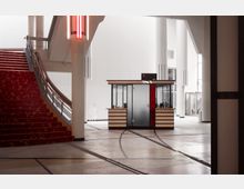 Großzügiger Innenraum mit moderner Architektur, einer roten Treppe mit gemustertem Teppich auf der linken Seite und einem kleinen, verglasten Kiosk in der Mitte. Helle Wände, hohe Decken und große weiße Säulen prägen die Umgebung.