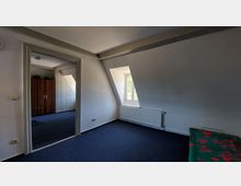 Ein Zimmer mit schräger Dachwand und kleinem Fenster, durch das Tageslicht einfällt. Sichtbar sind ein blauer Teppichboden, ein Heizkörper unter dem Fenster, sowie ein angrenzender Raum mit Garderobenschrank.