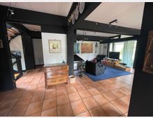 Wohnzimmer in einem Haus mit offener Gestaltung, gekennzeichnet durch schwarze Holzbalken, Terrakottafliesen und eine helle Holzdecke. Der Raum enthält ein Sofa, einen Fernseher, Regale, eine Kommode und große Fenster mit transparenten Vorhängen, die viel Tageslicht hereinlassen.