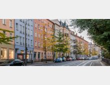 Straßenansicht mit mehrstöckigen Wohngebäuden in verschiedenen Pastellfarben, einer Baumreihe mit gelbem Herbstlaub und parkenden Autos; im Vordergrund eine Bushaltestelle und eine Straße mit Straßenbahnschienen.