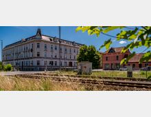 Straßenszene an einem Bahnübergang mit mehreren Gleisen im Vordergrund, hohem Gras und Signalanlagen. Dahinter stehen ein großes, historisches mehrstöckiges Eckgebäude mit dekorierter Fassade und Dachgauben sowie kleinere Häuser mit roten Ziegeldächern unter blauem Himmel.