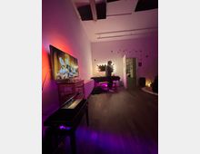 Long, loft-style room with white walls and a high ceiling, featuring purple ambient lighting, a large TV on a low cabinet, and a table with string lights near an open doorway. Acoustic panels are mounted on the ceiling, and the space has wood flooring with minimal furniture.