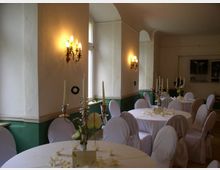 Ein eleganter Veranstaltungsraum mit weiß gedeckten Rundtischen und mit Stoffüberzügen versehenen Stühlen. Der Raum ist mit Wandleuchtern beleuchtet, verfügt über beige Wände und grüne Wandvertäfelungen sowie Fenster, die natürliches Licht einlassen.