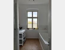 Bright bathroom with white half-height wall tiles and a wood floor, featuring a large arched window at the far end. A bathtub runs along the right wall, a small vanity sink sits on the left, and a glass shower partition is visible near the entrance.