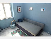 Kleines Zimmer mit blauen Wänden, einer grauen Schlafcouch, einem blauen Kissen und einer kleinen Lampe auf einem Beistelltisch. An der Wand hängen drei Bilder, und auf dem Boden liegt ein bunter Spielteppich neben einem großen Fenster.