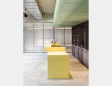 Minimalistisch gestalteter Innenraum mit einer zentralen gelben Theke, einer Decke mit grünen Glaspaneelen und einer Wand aus Regalelementen in dunkler Farbe. Der Raum hat durchsichtige Trennwände und sichtbare Lüftungsrohre an der Decke.