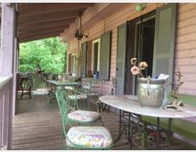 Veranda aus Holz mit floral verzierten Metallmöbeln und gemusterten Sitzpolstern, umgeben von grüner Vegetation. An der Holzwand des Gebäudes sind Fenster mit grauen Klappläden angebracht, auf einigen Tischen stehen dekorative Gegenstände.