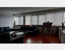 Sitzbereich in einem modern gestalteten Raum mit einer schwarzen Ledercouch, einem Glastisch und großen Fenstern mit vertikalen Jalousien. Rechts im Bild ist ein dekoratives Schild mit der Aufschrift „130 Jahre“ auf einem Ständer zu sehen, flankiert von Pflanzen und einem Schrank.