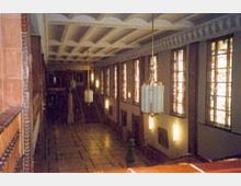 Innenansicht eines hohen Saals mit Kassettendecke, großen Fenstern, die seitlich angeordnet sind, und hängenden Kronleuchtern. Der Raum ist mit Holzelementen und geometrischen Verzierungen gestaltet.