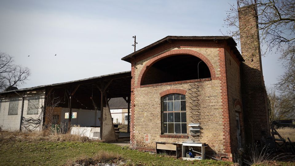 Backstein-Industriegebäude mit großem Rundbogen im Obergeschoss und hohem Schornstein, daneben eine offene überdachte Halle mit Stützen. Davor liegt eine verwilderte Rasenfläche mit Laub und vereinzelten Gegenständen; im Hintergrund sind kahle Bäume zu sehen.