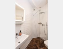 Modernes Badezimmer mit weiß gefliesten Wänden und dunklem Fliesenboden. Zu sehen sind ein Waschbecken mit Spiegel, eine gläserne Duschkabine mit Regenduschkopf und ein Handtuchheizkörper im Hintergrund.