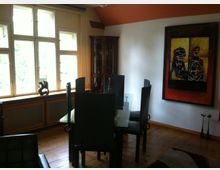 Esszimmer mit einem Glastisch und mehreren schwarzen Stühlen auf einem Holzboden. Große Fenster mit Holzrahmen lassen viel Tageslicht herein; an der Wand hängt ein farbenfrohes abstraktes Gemälde, daneben steht eine Vitrine mit Glasfront.